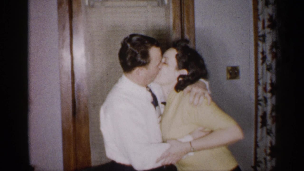 The adult couple remains in a close embrace beside the wooden doorway, with the man in a white shirt and tie holding the woman in a yellow top.