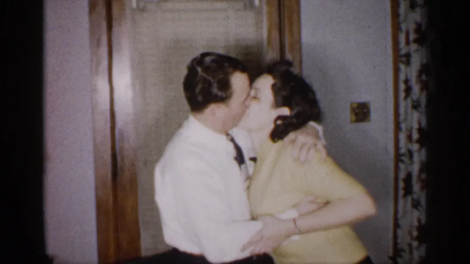The adult couple remains in a close embrace beside the wooden doorway, with the man in a white shirt and tie holding the woman in a yellow top.