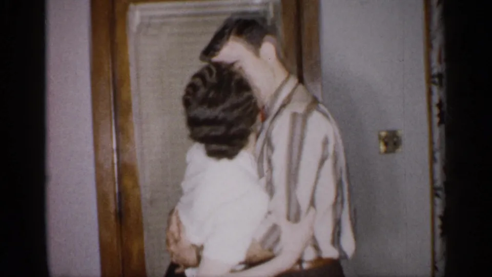 A second adult couple kisses in the domestic interior, with the man in a striped shirt holding a woman in a white blouse near the wooden doorframe.