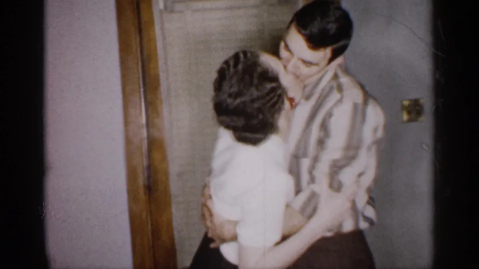 The second adult couple stays in a close embrace and kiss inside the Michigan domestic setting, with the striped shirt and white blouse visible against the doorway and wall.