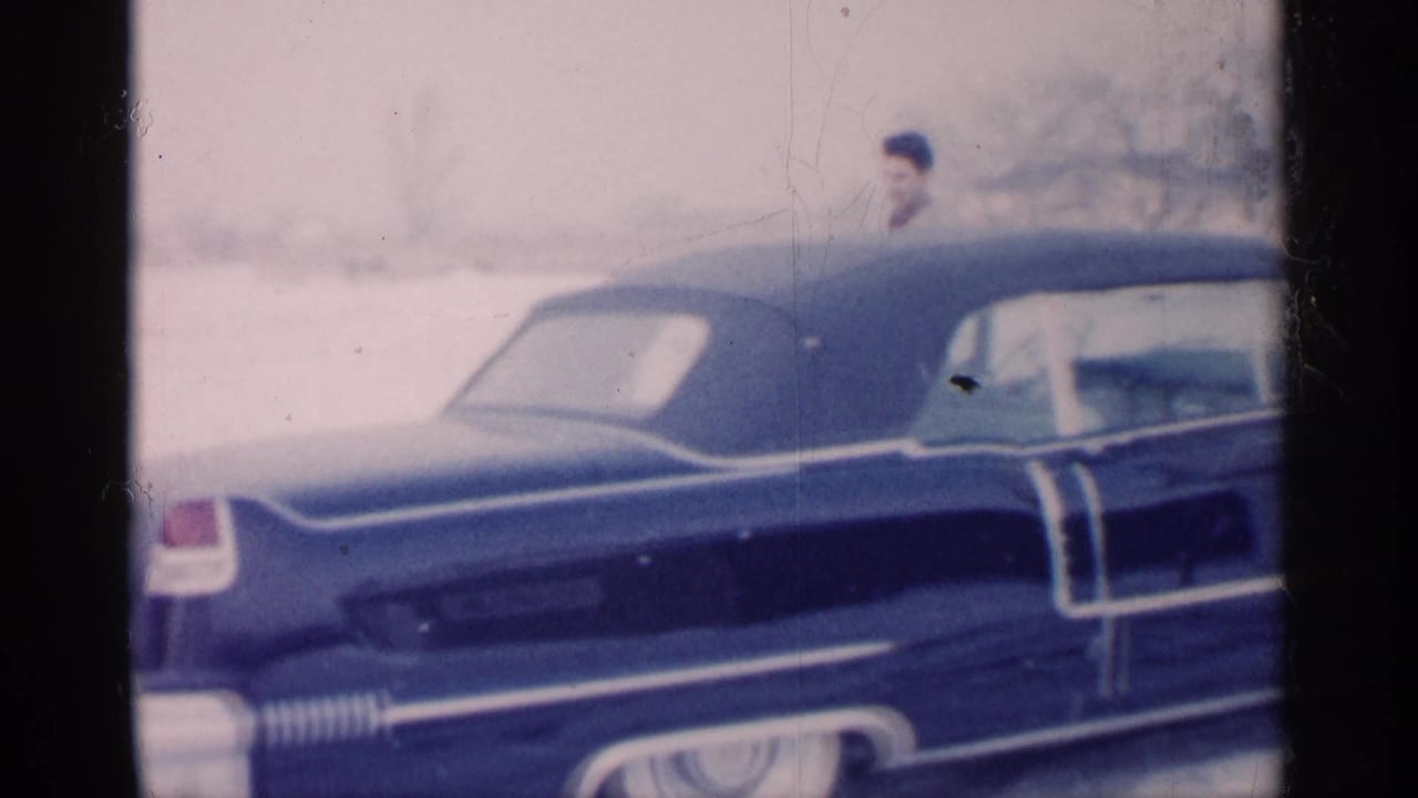 Blue vintage car in snow with a suited man partly visible behind it.