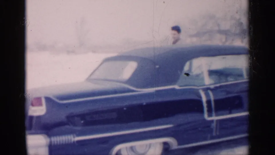 1959 Michigan home-movie frame showing a blue car with chrome trim and whitewall tire in snow, with a suited man partly visible.