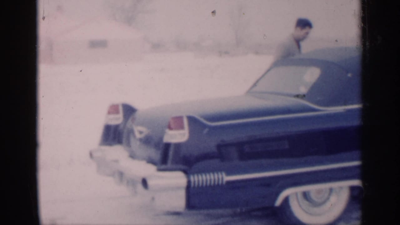Rear side of a blue vintage car in snow with a man near the roofline.