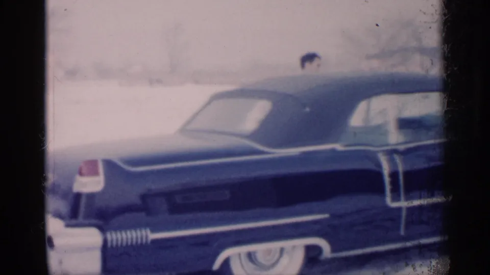 Soft 8mm frame from 1959 Michigan showing the side of a blue car in snow, with chrome detailing and a man partly visible.