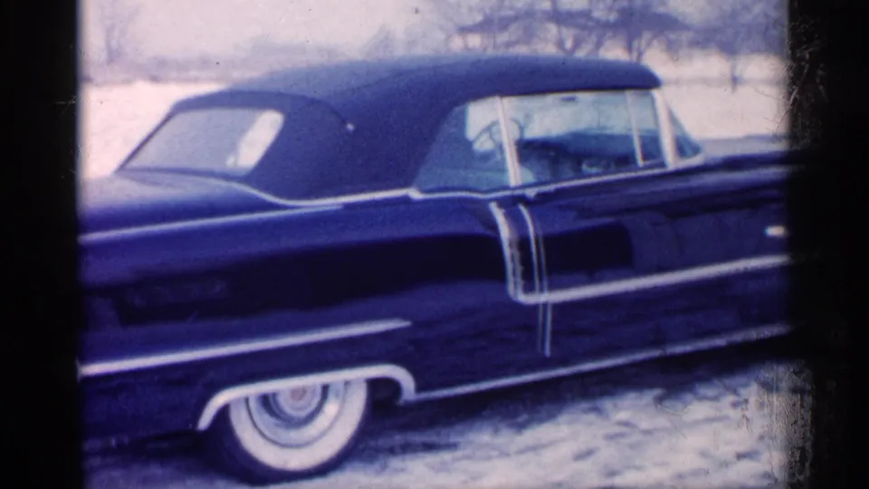 1959 Michigan home-movie frame showing a blue car in profile on snow, with chrome trim, whitewall tire, and empty-looking passenger area.