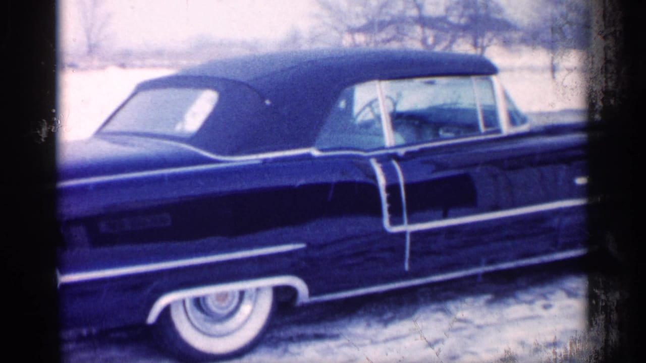 Michigan 1959 home-movie frame showing a dark blue convertible parked in snow, with chrome trim, whitewall tire, and winter trees.
