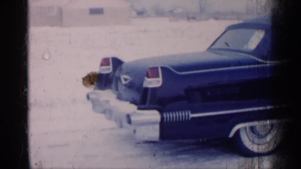 Michigan 1959 frame of the dark blue convertible from the rear side, showing snow, tailfin trim, and aged 8mm film texture.