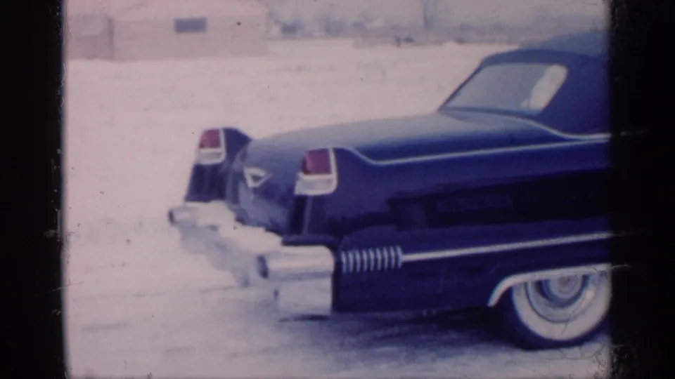 1959 Michigan home-movie frame showing a dark blue tailfinned convertible with chrome trim and whitewall tire in snow.