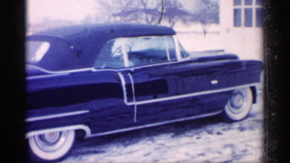1959 Michigan home-movie frame with a closer side view of the dark blue convertible, whitewall tires and snowy yard visible.
