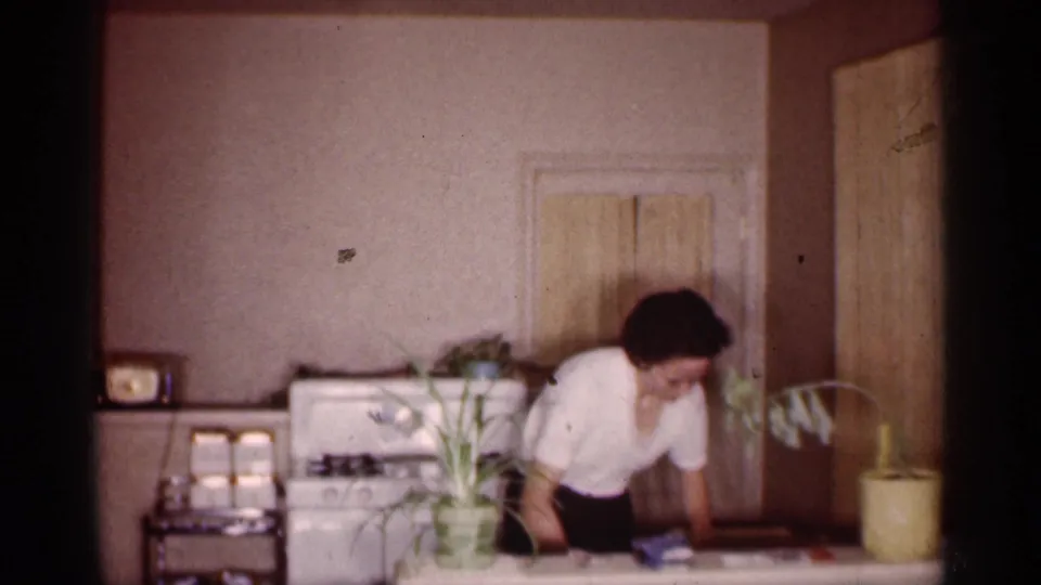 1959 Michigan home-movie frame showing a woman in a kitchen beside a counter, stove, potted plants and domestic furnishings.