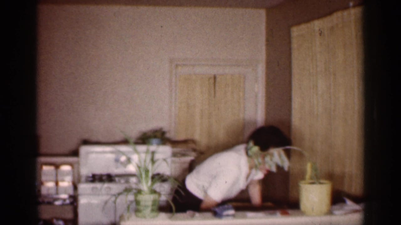 Woman leans over a kitchen table near a white stove and potted plants.