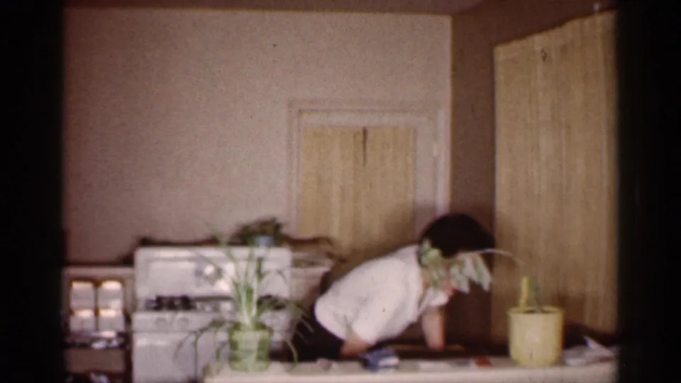 In a 1959 Michigan home-movie frame, a woman bends over a kitchen table beside a white stove, potted plants, and wood-paneled doors.