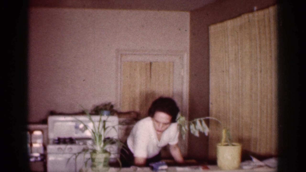 Woman bends over the kitchen table with plants and a stove behind her.