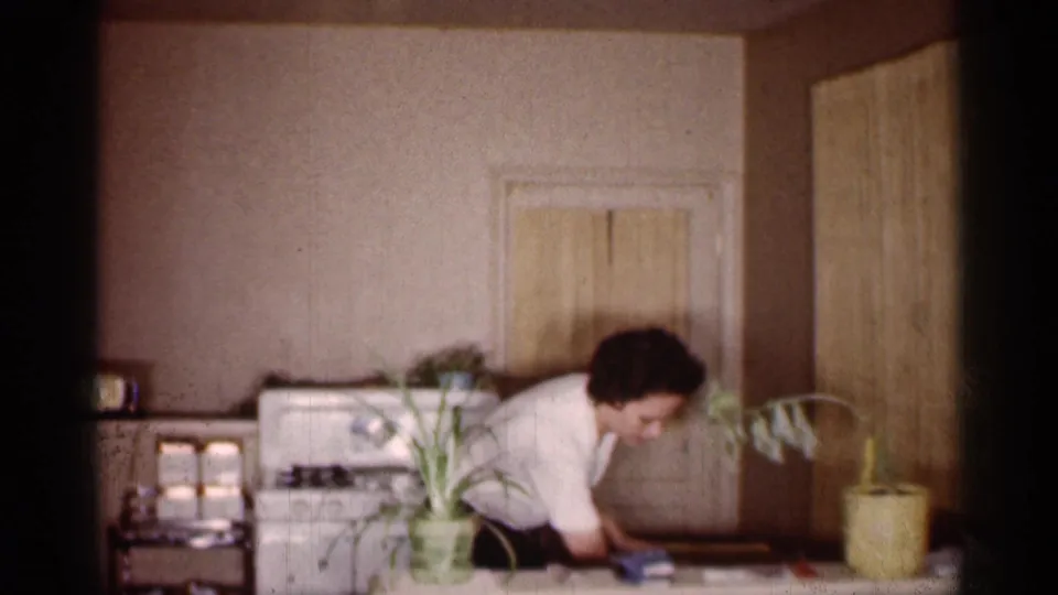 The home-movie frame shows a woman reaching across a kitchen table, partly obscured by potted plants, with a white stove behind her.