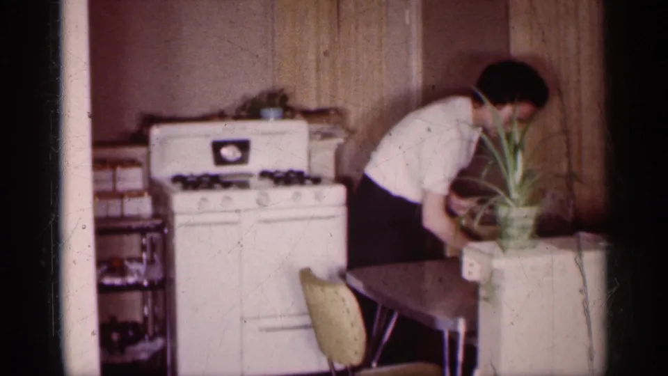 A woman stands at the side of a kitchen table in the 1959 Michigan home movie, with a white stove, potted plant, yellow chair, and wood paneling visible.