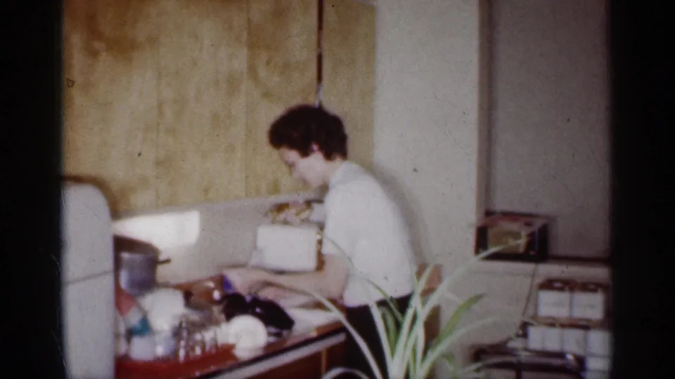 In 1959 Michigan home-movie footage, a woman stands at a kitchen sink near a white refrigerator, glassware, and a potted fern.