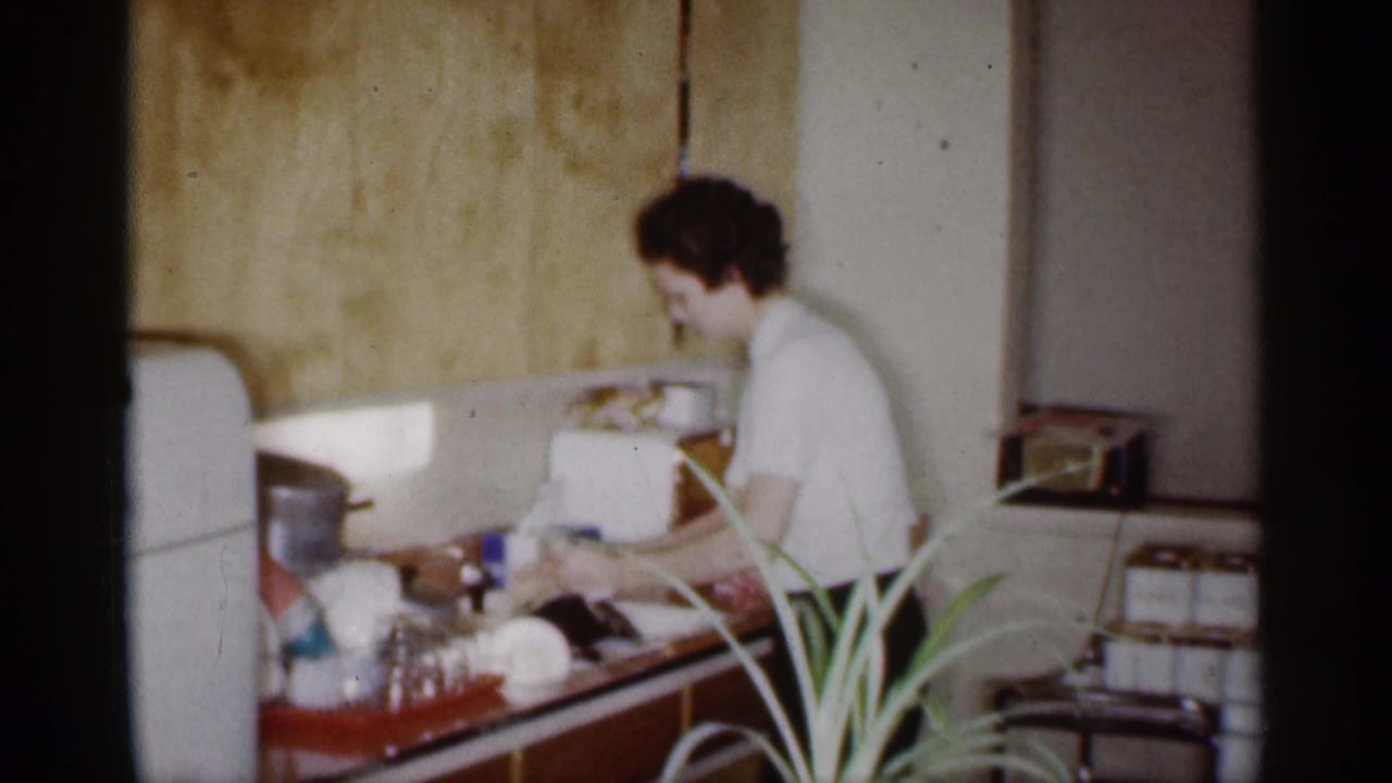 Woman continues washing dishes at a counter with glassware and a small radio nearby.