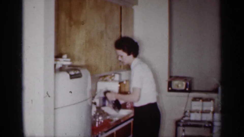 A woman in a white top and dark skirt leans over the sink in a 1959 Michigan kitchen, with dishes and glassware on the counter.