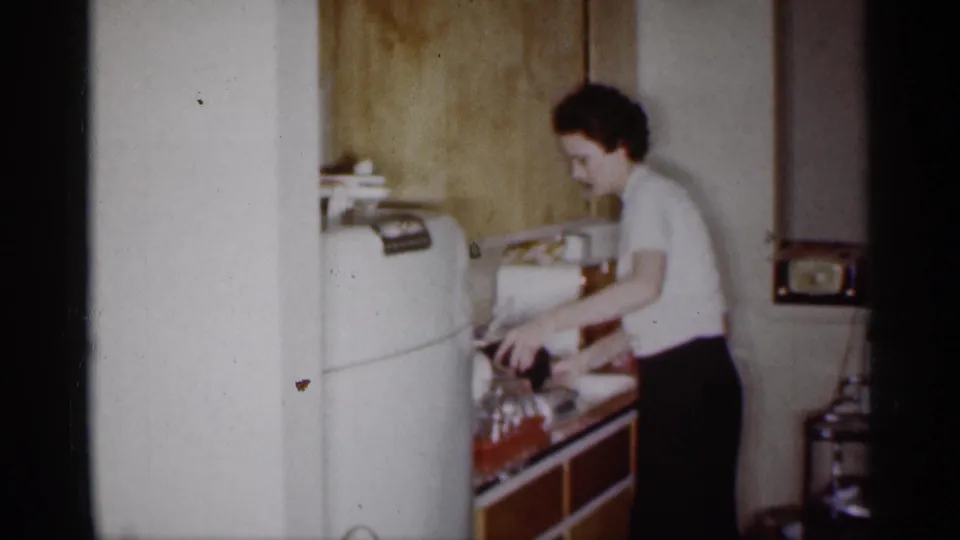 The woman reaches along the counter in the private Michigan kitchen, with the refrigerator, sink area, glassware, and small radio visible.