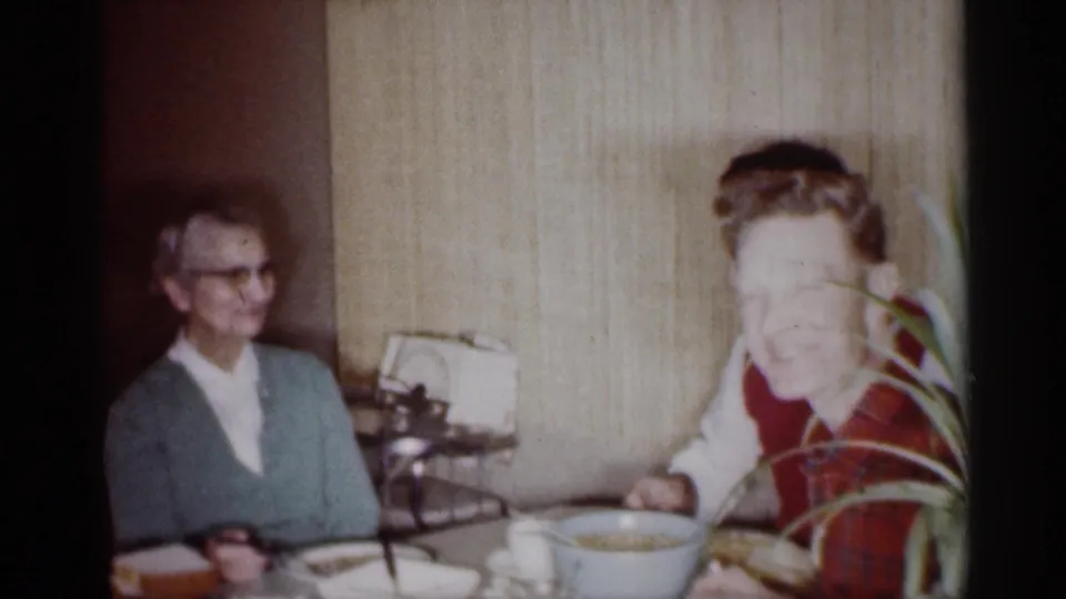 1959 Michigan home-movie frame showing an older woman and a young boy seated at a dining table in a private home.