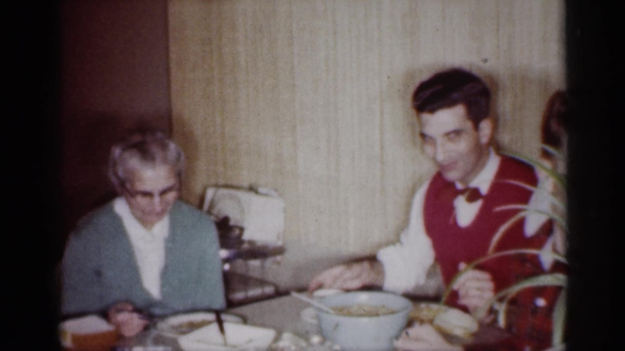 Older woman looks down while a young man in a red vest sits beside the table.