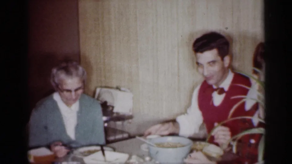 1959 Michigan home-movie frame with an older woman and a young man in a red vest seated at a dining table with bowls and cups.