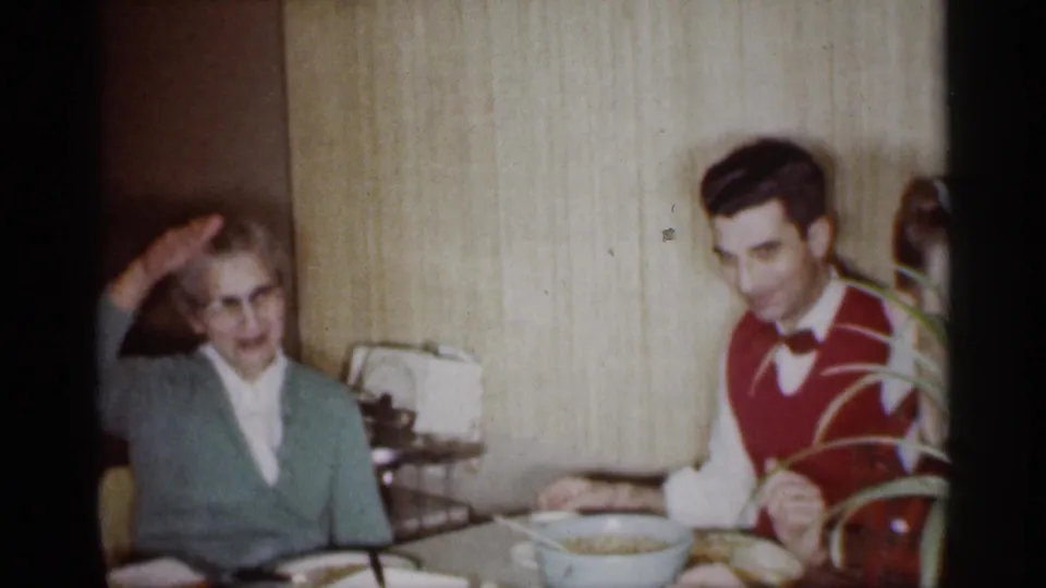 1959 Michigan home-movie frame showing an older woman gesturing at a dining table as a young man in a red vest looks toward the camera.