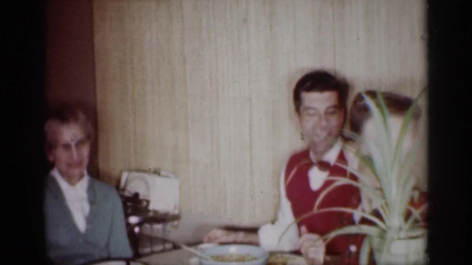 1959 Michigan home-movie frame with an older woman and smiling young man at a dining table, partly framed by a houseplant.