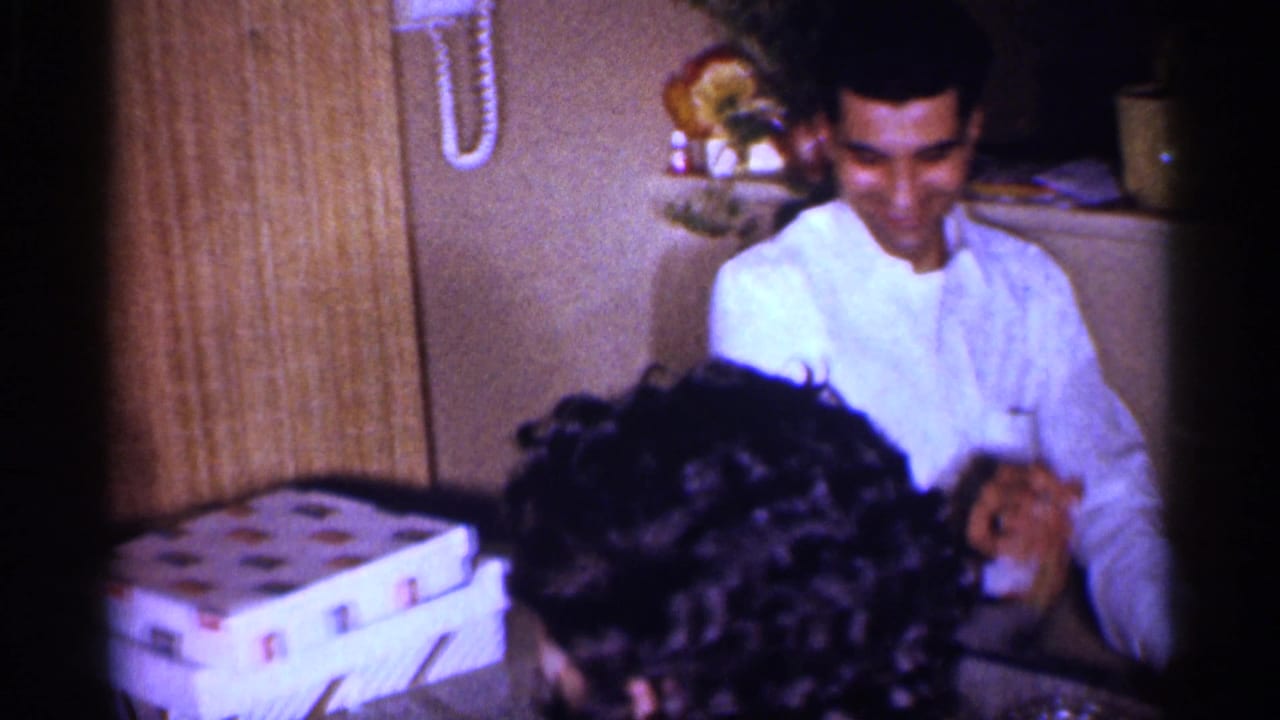Young man in a white shirt smiles at a table while a dark-haired woman is seen from behind.