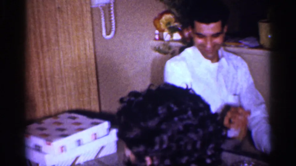 1959 Michigan home-movie frame showing a smiling young man at an indoor family table with a woman in the foreground.