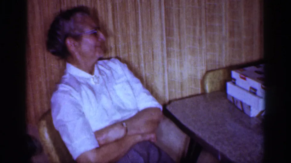 1959 Michigan frame of an older woman seated with arms folded near a table in a wood-paneled family room.