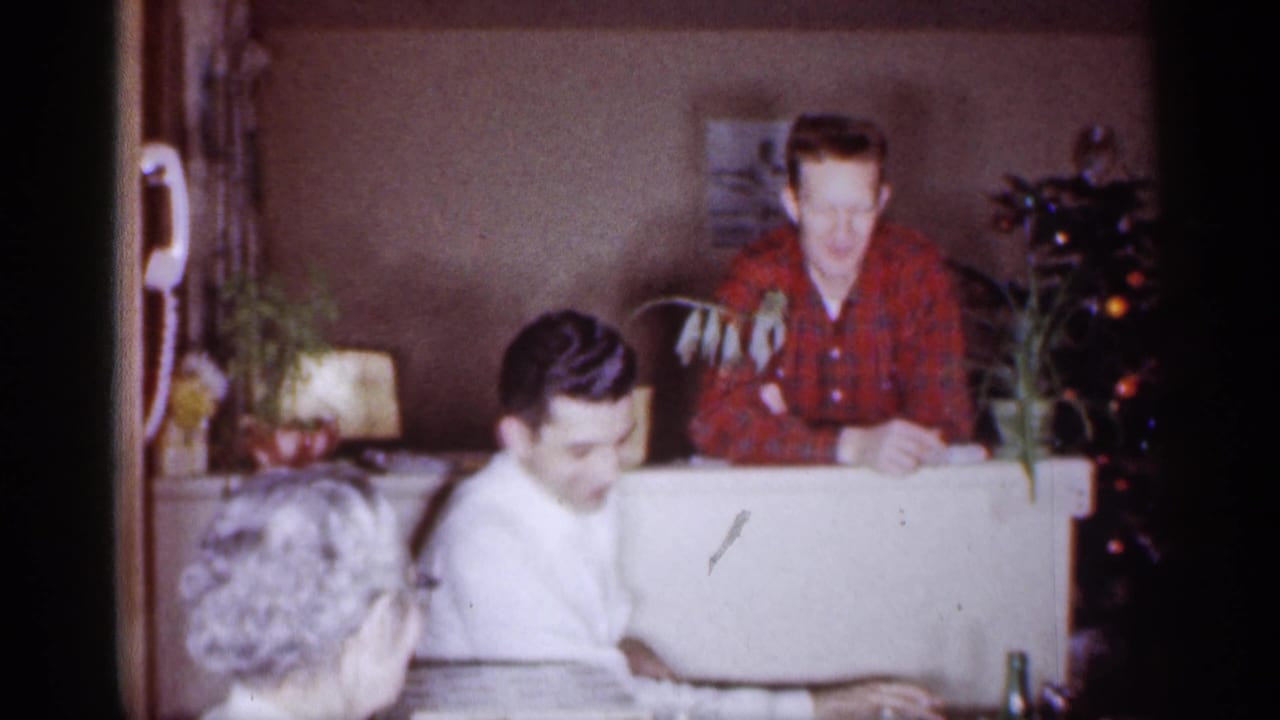 1959 Michigan home-movie frame with adults gathered indoors near plants and a Christmas tree, with a man in red plaid at the back.