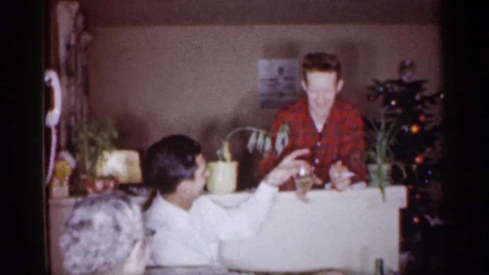 1959 Michigan home-movie frame showing a man in white handing a glass to a man in red plaid during an indoor holiday gathering.