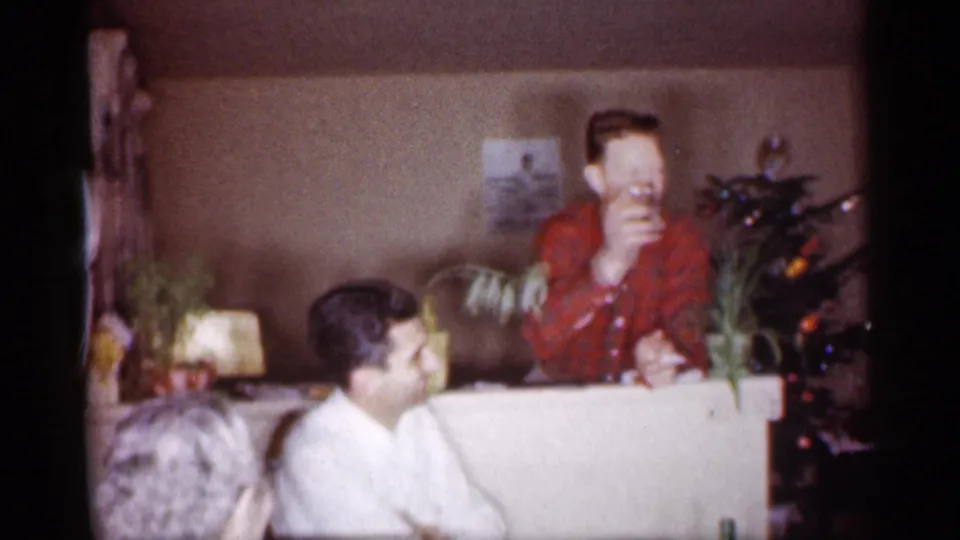 1959 Michigan home-movie frame with a man in red plaid raising a drink beside a Christmas tree as another adult looks on.