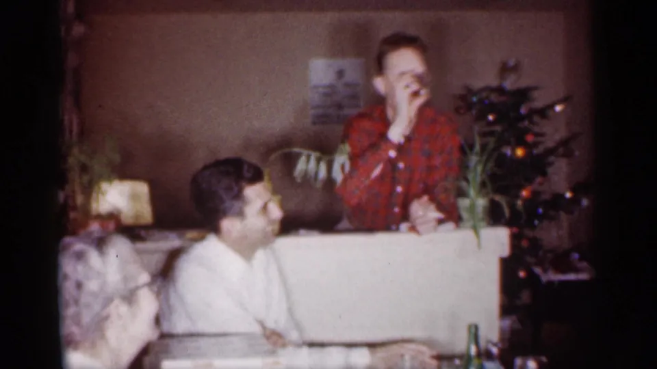 1959 Michigan home-movie frame showing a man in red plaid taking a sip beside a Christmas tree in a private interior.