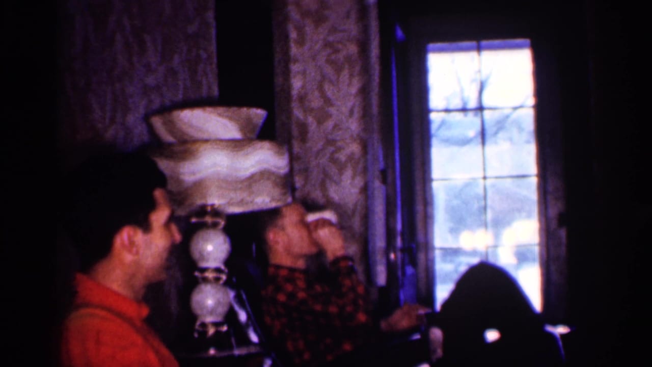 Two adult men sit in a dim living room near a tiered lamp and window.