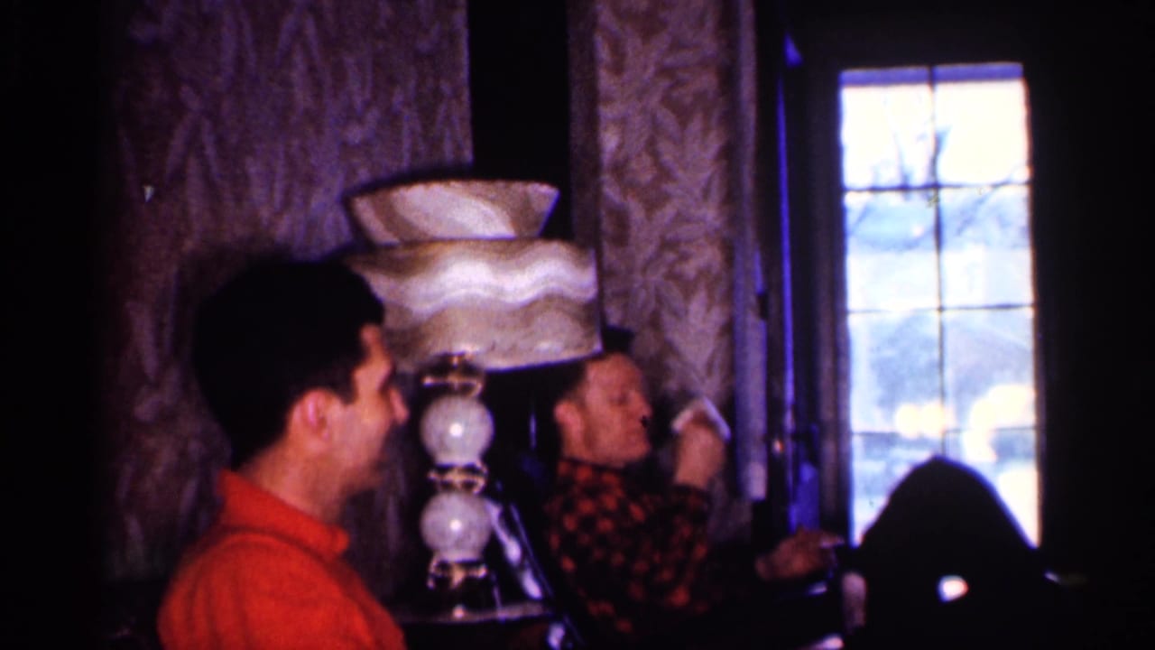Two men sit beside a vintage lamp as one raises a hand near his face.