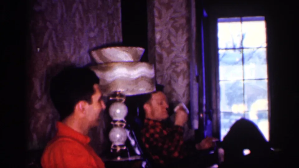 Two adult men in a Michigan living room sit beside a tiered lamp, with one man in plaid lifting his hand near his face.