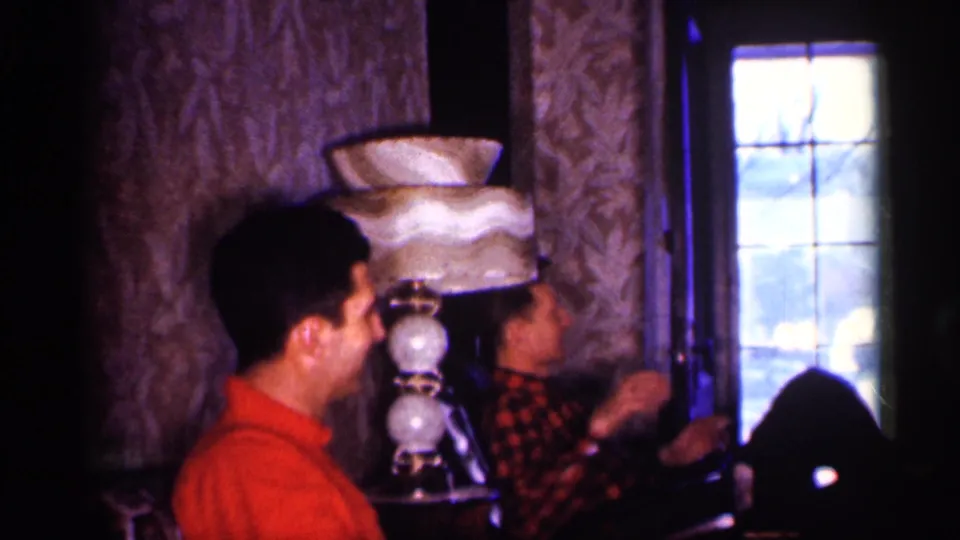 The camera frames two adult men seated close together in a 1959 Michigan living room with a tiered lamp and bright window.