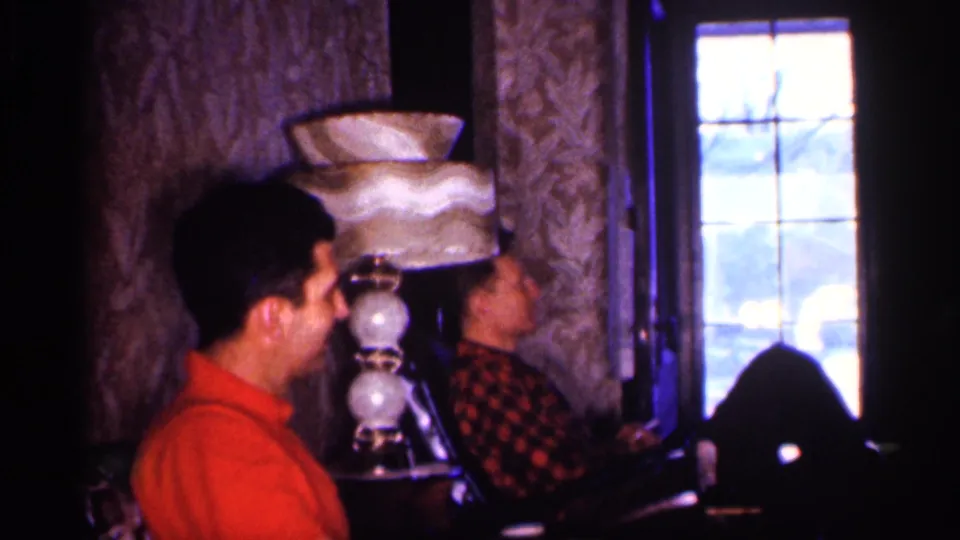 In the living-room scene, the man in red sits in profile while the man in plaid reclines near a tiered lamp and window.