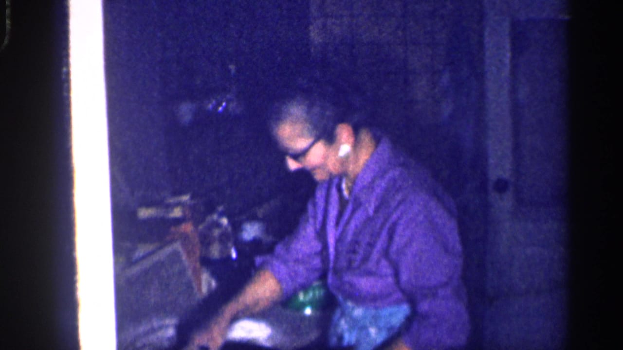 Older woman in glasses works at a dim kitchen counter.