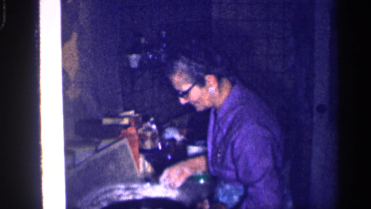 The older woman continues working at the kitchen counter, with a green bottle and cabinet surfaces visible in the dim 1959 Michigan interior.