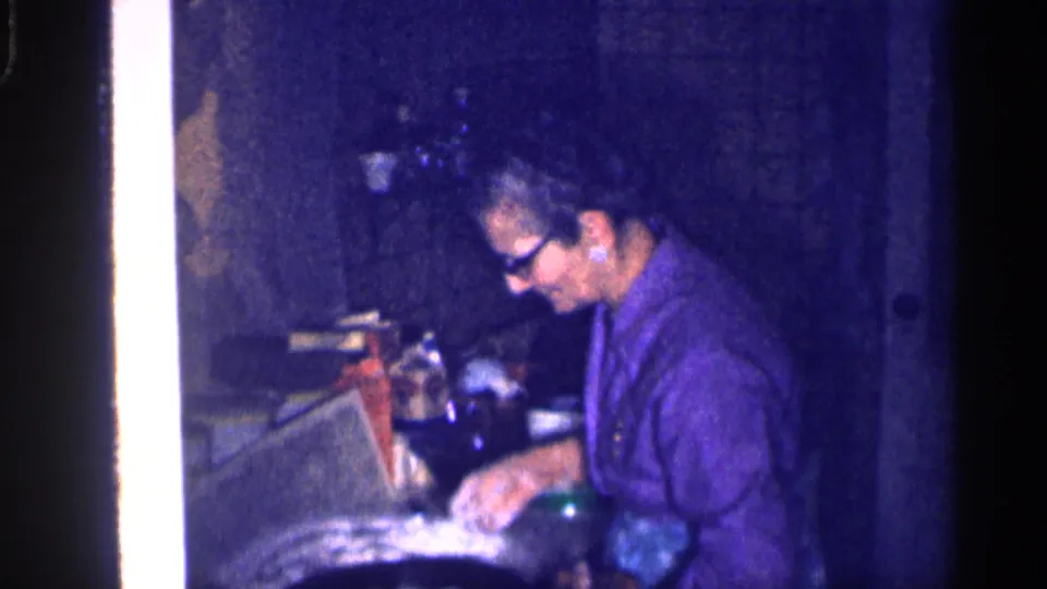 The older woman continues working at the kitchen counter, with a green bottle and cabinet surfaces visible in the dim 1959 Michigan interior.