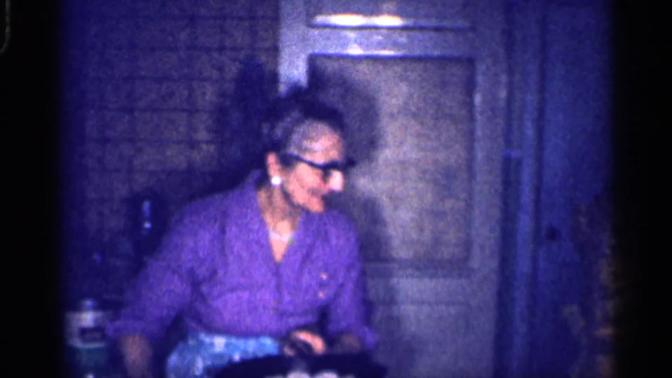 The woman turns near paneled cabinetry and a brick or tile wall surface inside the dim Michigan kitchen recorded on 8mm film.