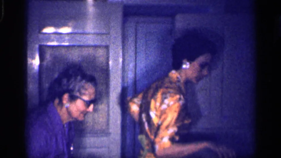 A second woman in a patterned dress enters the dim kitchen as the older woman in purple remains near the cabinetry.