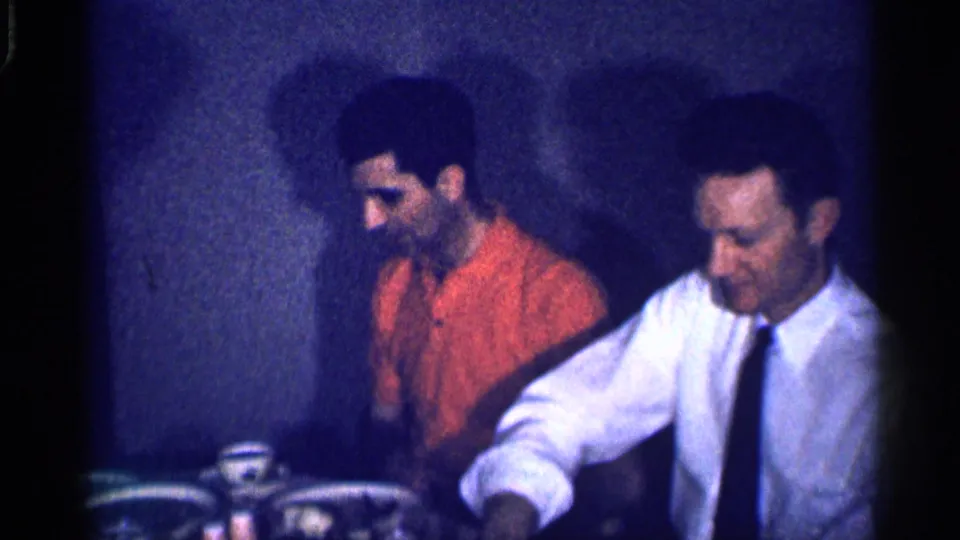 In the 1959 Michigan home movie, two adult men sit at a table, one in an orange shirt and one in a white shirt with a dark tie, with cups and plates nearby.