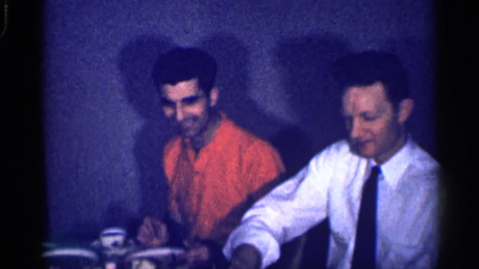 Two adult men remain seated at the table in the 1959 Michigan footage, with the man in orange smiling while cups, plates, and food sit in front of them.