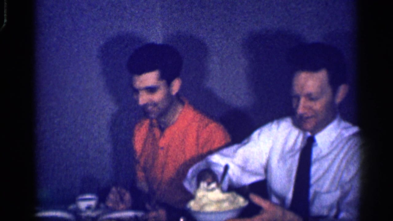 Two adult men sit at a table as a bowl of mashed potatoes is held near them.