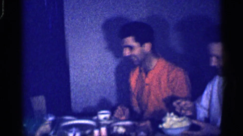 The home movie frames a smiling man in an orange shirt at a Michigan indoor meal, with tableware and food in front of him.