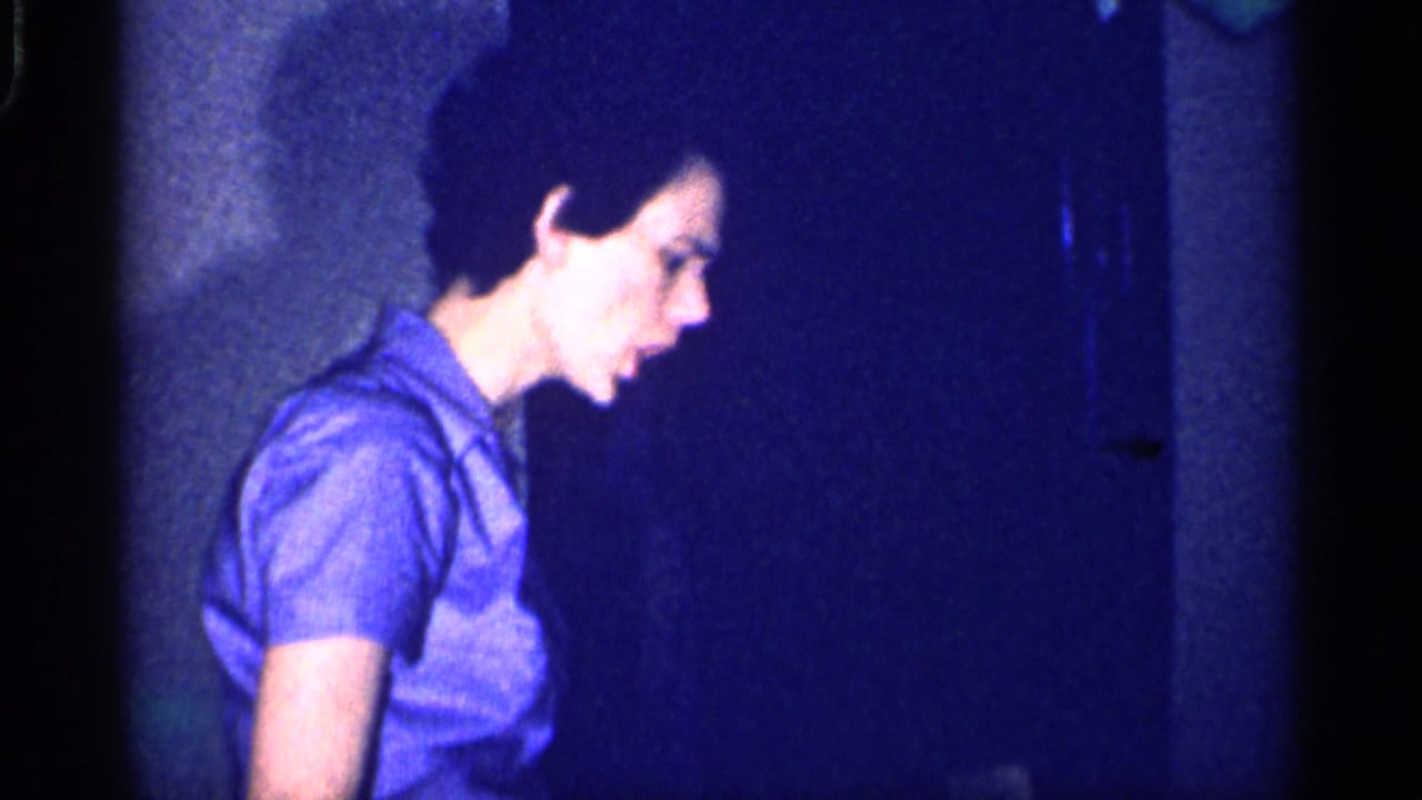 Woman in a blue dress stands in a dim interior near a wall or doorway.
